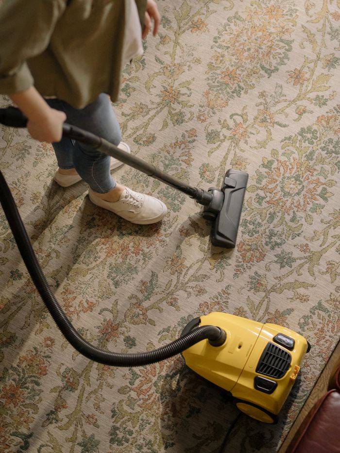 A person is cleaning a floral rug indoors with a yellow vacuum cleaner.